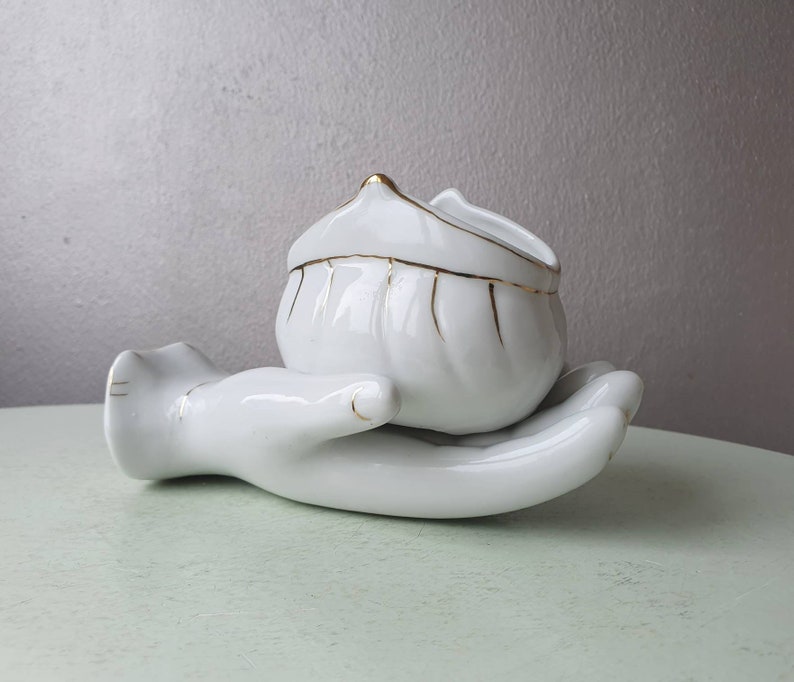 May include: White ceramic hand-shaped dish with a white ceramic bowl with a gold trim sitting in the palm of the hand.