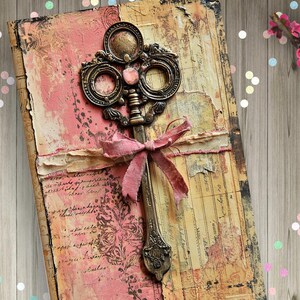 May include: A rectangular journal with a distressed cover featuring shades of pink, cream, and brown. The cover displays a large, ornate, metallic bronze-colored key embellishment tied with a frayed pink fabric ribbon across the center.