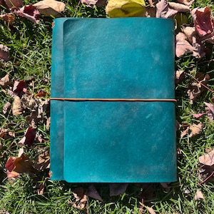 May include: A teal leather journal with a brown leather strap wrapped around it. The journal is lying on a bed of green grass and fallen autumn leaves. The journal is closed and appears to be new.