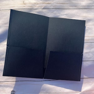 May include: A black, open presentation folder with two interior pockets. The folder is made of a matte material and is displayed open on a light-colored wooden surface. The folder's design is simple and professional.