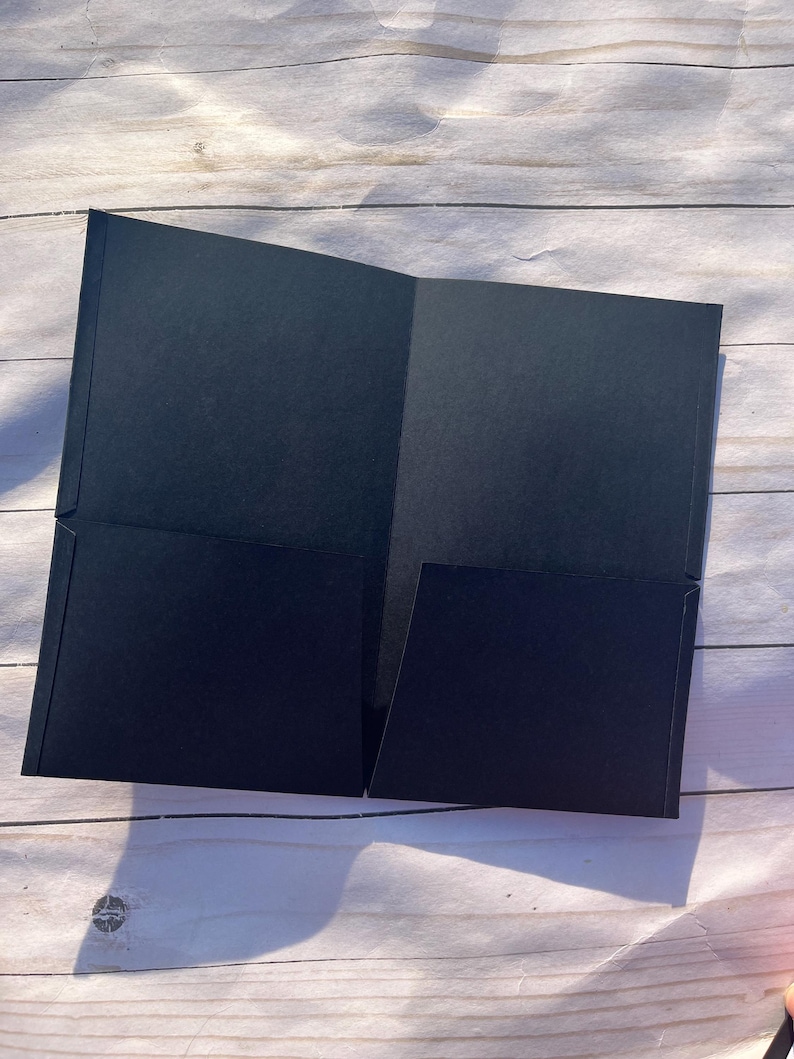 Peut inclure: Un porte-documents noir ouvert, r&eacute;v&eacute;lant deux poches int&eacute;rieures. Le dossier est fait d'un mat&eacute;riau mat et est pr&eacute;sent&eacute; ouvert sur une surface en bois de couleur claire. Le design du dossier est simple et professionnel.