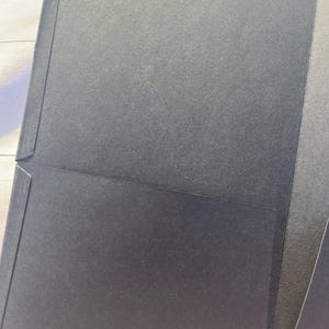 Peut inclure: Un porte-documents noir avec une surface textur&eacute;e. Le dossier a une poche &agrave; l'int&eacute;rieur et est ouvert, r&eacute;v&eacute;lant l'int&eacute;rieur. Le dossier est fait d'un mat&eacute;riau en papier &eacute;pais et durable.