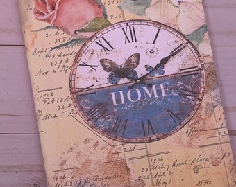 May include: A vintage-style clock with a blue and white face and roman numerals. The clock has a gold-colored frame and the words "HOME is where the heart is" printed on the face. The clock is surrounded by a floral design with a pink rose and a white flower.
