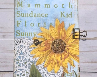 May include: A blue and white journal with a sunflower, a bee, and lace design. The text on the cover reads "Suntastic Yellow Mammoth Sundance Kid Floris Sunny".