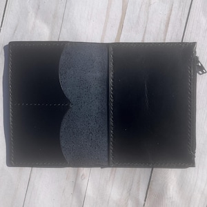 May include: Open black leather wallet with a zippered compartment and multiple card slots. The wallet is shown open, revealing a textured interior and contrasting stitching. The design is simple and functional.