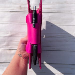 May include: A bright pink leather pen holder with a black S.GEL pen inside. The pen holder is held upright, showcasing its design and the pen's details. The pen is black with silver accents and the text "S.GEL" printed on the side.