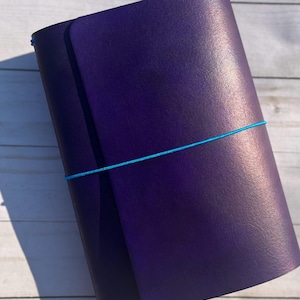 May include: A dark purple leather journal with a light blue elastic closure. The journal is closed and has a textured surface. The journal is a notebook or diary.