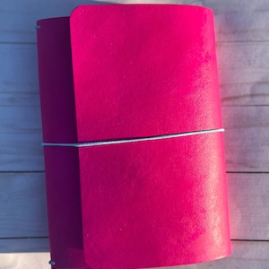 May include: A bright pink journal with a light blue elastic closure. The journal is closed and has a smooth, matte finish. The cover wraps around the journal and is secured with a light blue elastic band.
