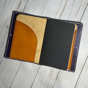 May include: A purple leather notebook cover with a tan leather pocket and a black notebook insert.