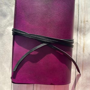 May include: A handmade journal with a deep purple cover and a black leather tie closure. The journal's cover has a gradient effect, transitioning from a lighter shade at the top to a darker shade at the bottom. The journal is closed with a black leather tie.