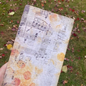 May include: A vintage-style notebook with a distressed white cover featuring a musical score and orange and yellow circular patterns.