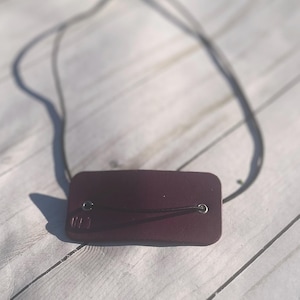 May include: A rectangular, burgundy-colored leather tag with a dark cord attached. The tag has two metal grommets and a small embossed detail on the lower left corner. The cord is looped through the grommets.