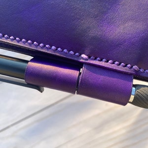 Könnte beinhalten: Nahaufnahme eines violetten Ledernotizbuchs mit Stifthalter. Das Notizbuch hat einen genähten Rand und der Stifthalter ist aus dem gleichen Leder.