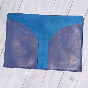Puede incluir: Una cartera de cuero hecha a mano en tonos azules. La cartera presenta una sección central de cuero azul brillante, flanqueada por dos bolsillos curvos de un cuero azul-púrpura más oscuro. Los bordes están cosidos con hilo en contraste.