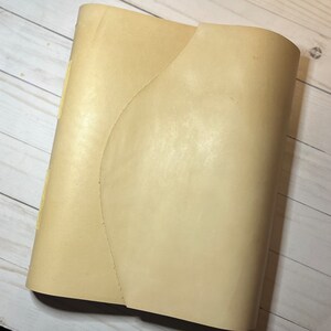 May include: A cream-colored leather journal with a unique curved spine. The journal is closed and has a smooth, textured surface.