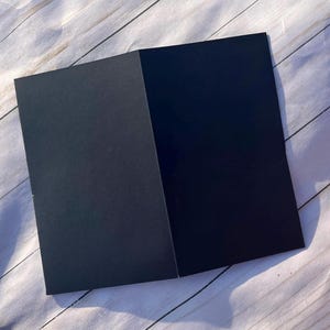 Peut inclure: Une carte noire pli&eacute;e, ouverte pour r&eacute;v&eacute;ler un int&eacute;rieur noir plus fonc&eacute;. La carte est positionn&eacute;e en diagonale sur une surface claire &agrave; grain de bois, cr&eacute;ant un contraste entre la carte sombre et le fond clair. Les bords de la carte sont nets et propres.