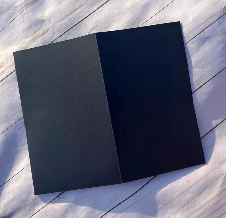 Peut inclure: Une carte noire pli&eacute;e, ouverte pour r&eacute;v&eacute;ler un int&eacute;rieur noir plus fonc&eacute;. La carte est positionn&eacute;e en diagonale sur une surface claire &agrave; grain de bois, cr&eacute;ant un contraste entre la carte sombre et le fond clair. Les bords de la carte sont nets et propres.