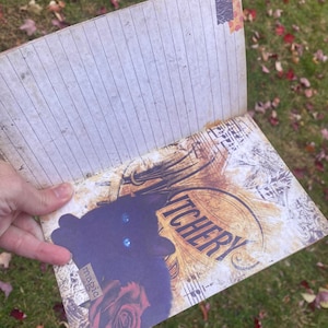 May include: A black cat with blue eyes is featured on the cover of a journal with a vintage design. The journal has a lined interior and the cover reads "Witchery" with the word "magic" written on a tag.