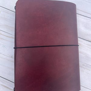 May include: A burgundy leather journal with a black elastic closure. The journal is rectangular and has a smooth, textured surface. The cover is a deep, rich burgundy color.