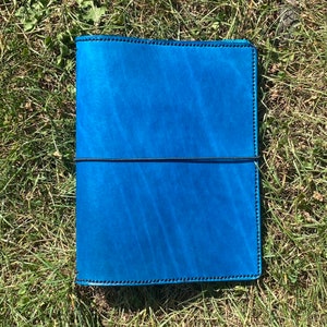 May include: A rectangular blue leather notebook cover featuring a smooth, vibrant finish with visible grain texture. The cover is secured by a thin black elastic band wrapped horizontally around the center and is finished with dark stitching along the edges.