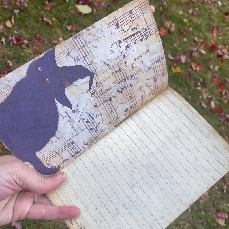 May include: A black cat with a witch's hat is printed on the front cover of a blank journal with lined pages. The cover is decorated with a vintage music sheet background.