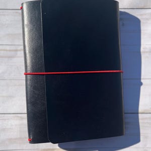 May include: A black leather journal with a red elastic closure. The journal is closed and has a smooth, matte finish. The journal is rectangular and appears to be new.