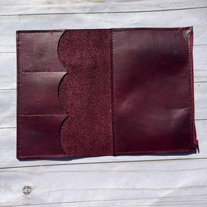 Può includere: Un portafoglio in pelle bordeaux, aperto per mostrare tasche interne e uno scomparto centrale. Il portafoglio ha una chiusura con cerniera ed è posto su uno sfondo chiaro con venature del legno. La pelle ha un aspetto ricco e strutturato.