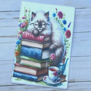 May include: A white cat with glasses is sleeping on a stack of colorful books. The cat is surrounded by flowers and a teacup with a saucer.