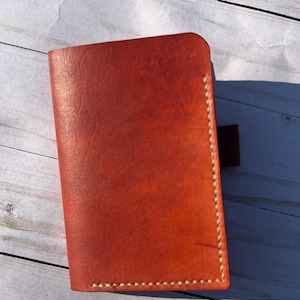 May include: A handcrafted, orange-red leather notebook cover. The cover is rectangular with rounded corners and a stitched edge. A small, matching leather tab is attached to the right side. The leather has a natural, textured appearance.