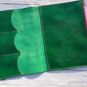 May include: Open green leather journal with a pink zipper and a green tab closure. The interior features a lighter green pocket design with scalloped edges and stitched details. The journal is on a white wooden surface.