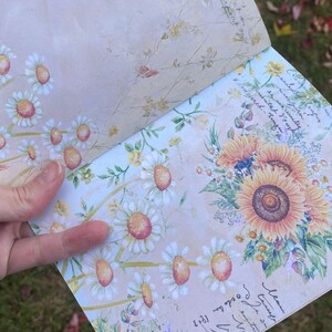 Op de afbeelding: Een lichtblauw en beige dagboek met een bloemenontwerp. De omslag heeft witte madeliefjes en gele zonnebloemen. De tekst "Mean Girls" staat in de rechterbenedenhoek van het dagboek.