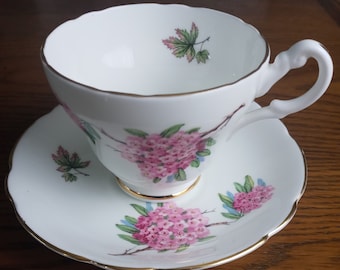 May include: A white teacup and saucer with a floral design. The cup and saucer have a gold trim and feature pink flowers and green leaves.