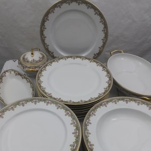 Może przedstawiać: Zestaw białej porcelanowej zastawy stołowej z pozłacanym rantem i kwiatowym wzorem. Zestaw zawiera dużą paterę, miskę do zupy, cukiernicę i stos talerzy obiadowych.