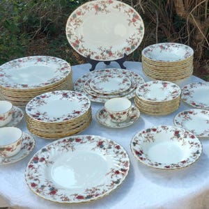 May include: A complete vintage floral china set, including plates, bowls, cups, saucers, and an oval platter. The white porcelain features a red, green, and gold floral pattern. The set is arranged on a white tablecloth.