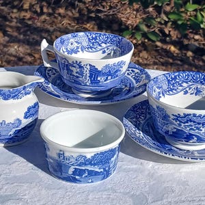 May include: A blue and white porcelain tea set featuring a scenic landscape design. The set includes two teacups with saucers, a creamer, and a small bowl. The teacups and creamer have handles. The set is placed on a white tablecloth.