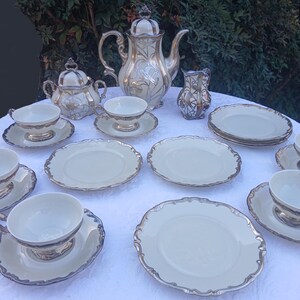 May include: A vintage tea set with a cream and gold color scheme. The set includes a teapot, sugar bowl, creamer, cups, saucers, and plates. The edges of the plates and cups are trimmed with gold. The set is arranged on a white tablecloth.