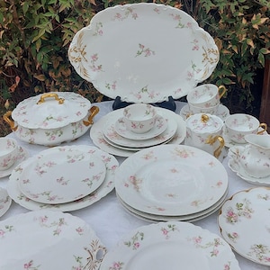 Puede incluir: Un juego completo de vajilla antigua de porcelana. El juego incluye fuentes, platos, tazas, platillos, azucarero, cremera y una fuente cubierta. Cada pieza es blanca con un delicado estampado floral rosa y ribete dorado.