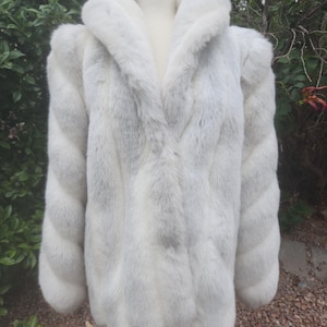 May include: A white faux fur coat featuring a thick, plush texture and a large, wide collar. The coat has long sleeves with a distinct vertical, segmented pattern throughout the body and arms, creating a soft, voluminous appearance.