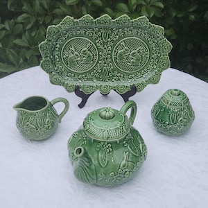 Op de afbeelding: Een saliegroene keramische theeset bestaande uit een theepot, een melkkannetje, een suikerpot en een ovaal dienblad. Elk stuk is versierd met reliëffiguren van konijnen, bladeren en gestructureerde rasterpatronen over het oppervlak.