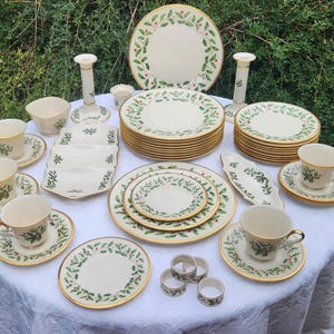 May include: A large set of cream-colored ceramic dinnerware featuring a green holly leaf and red berry pattern with gold trim. The collection includes dinner plates, salad plates, teacups, saucers, candlesticks, napkin rings, and serving dishes.