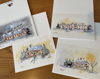 以下が含まれることがあります： 水彩画のグリーティングカード。冬の風景が描かれており、雪の家、サンタのそり、ヘラジカなどが描かれています。カードは木製の表面に置かれています。