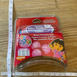 Puede incluir: Paquete protector de casco para niños pequeños Dora la Exploradora con rodilleras y coderas, y rodilleras y coderas adicionales. El paquete es rojo y rosa con una imagen de Dora en la parte delantera. El embalaje dice "Helmet Protective Pack" y "Offre Protection Casque".
