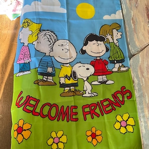 Może przedstawiać: Prostokątna flaga z postaciami z Peanuts na tle błękitnego nieba z żółtym słońcem i białymi chmurami. Flaga ma czerwony napis "WELCOME FRIENDS". Na dole znajdują się żółte i pomarańczowe kwiaty.