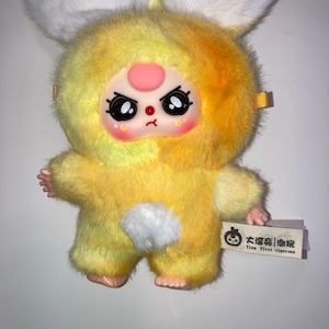 May include: A plush toy in the shape of a yellow bunny with a grumpy face. The toy has large white ears, a pink nose, and a white fluffy tail. A tag with Chinese characters and the text "Tivu First rigorous" is attached.