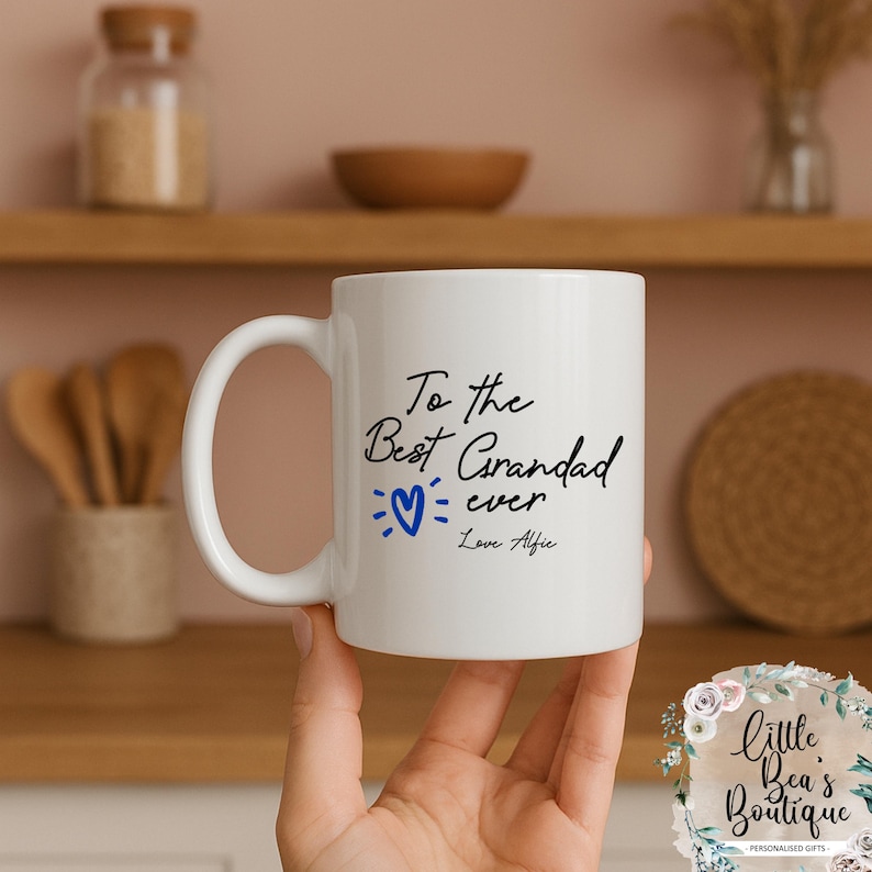 May include: White ceramic mug with the text "To the Best Grandad ever" in black script, a blue heart, and "Love Alfie" in a handwritten font. The mug has a curved handle.
