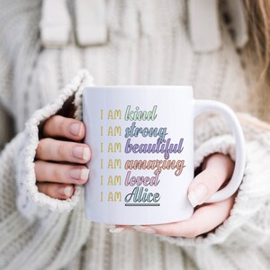 Op de afbeelding: Een witte keramische mok met een kleurrijk ontwerp met de tekst "Ik ben aardig, ik ben sterk, ik ben mooi, ik ben geweldig, ik ben geliefd, ik ben Alice".