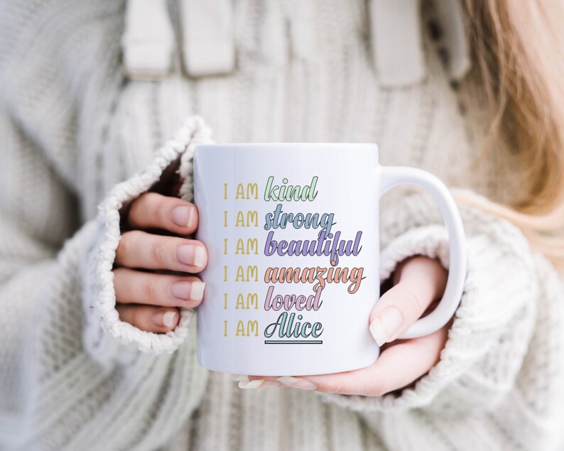 Op de afbeelding: Een witte keramische mok met een kleurrijk ontwerp met de tekst "Ik ben aardig, ik ben sterk, ik ben mooi, ik ben geweldig, ik ben geliefd, ik ben Alice".