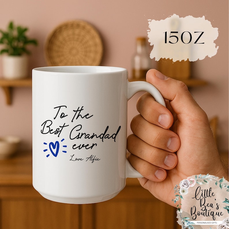 May include: White ceramic mug with the text "To the Best Grandad ever Love Alfie" in black script, a blue heart, and a 15 oz capacity. The mug has a white handle and is held by a person.