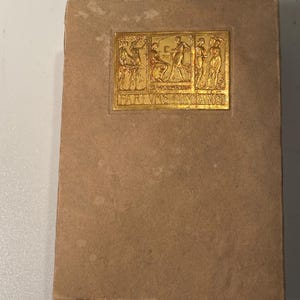 May include: A rectangular, tan-colored book with a gold-colored decorative plaque. The plaque features a relief of figures and the text "PARE VAS FONTANIS". The book has a worn appearance.