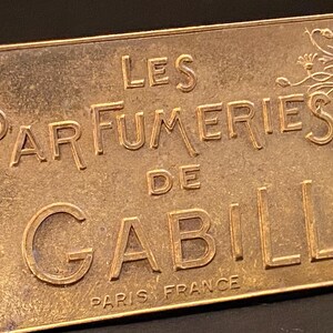 Peut inclure: Une plaque en métal dorée avec le texte "Les Parfumeries de Gabilla Paris France". La plaque a un motif floral dans le coin supérieur droit.